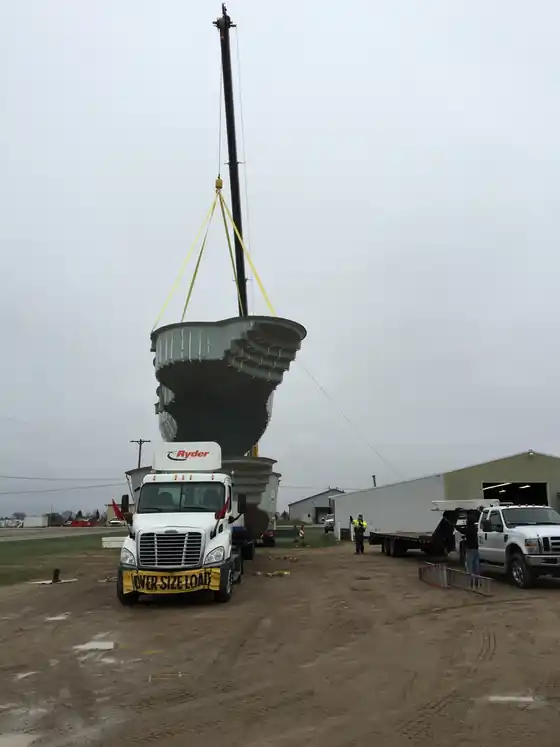 A crane lifting a fiberglass pool shell off a flatbed truck beside the lake