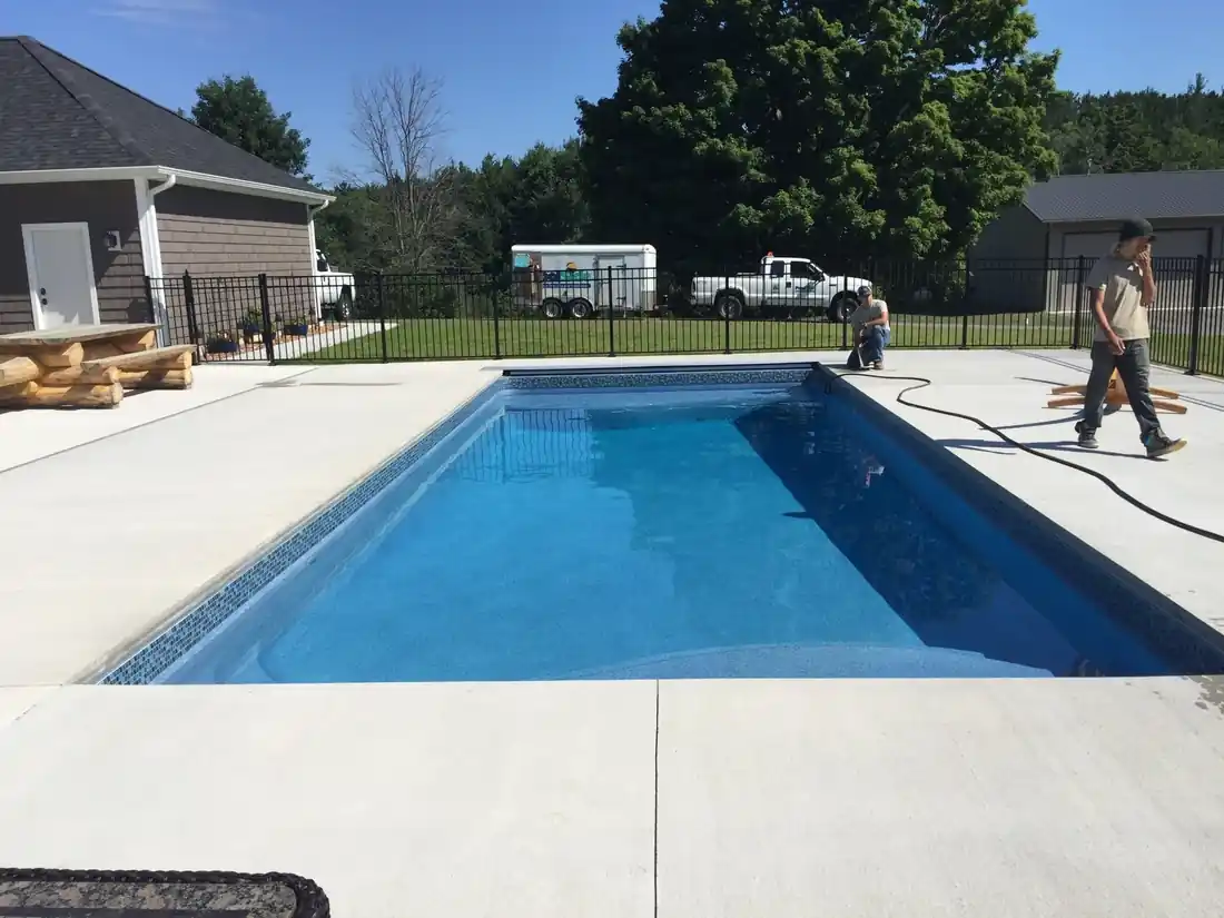 A finished rectangular fiberglass pool with an exposed aggregate deck and a crew member walking the edge